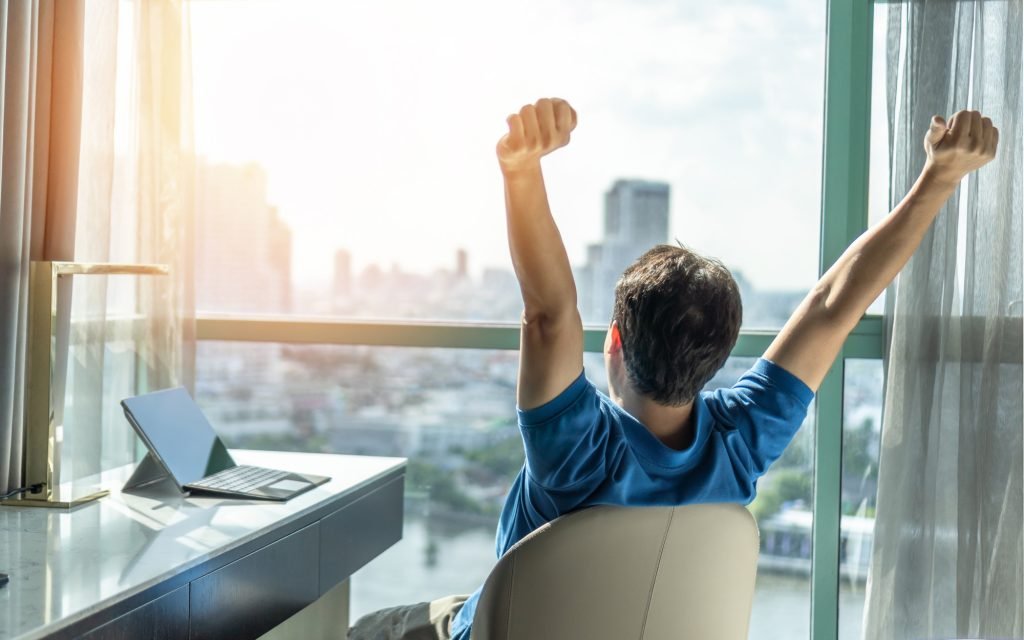 Business achievement concept with happy businessman relaxing in office or hotel room, resting and raising fists with ambition looking forward to city building urban scene through glass window
